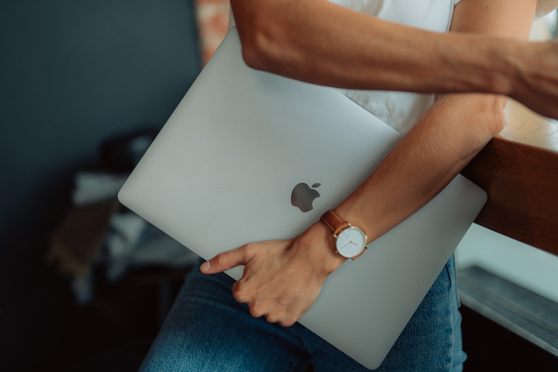 A person with a watch, holds their closed Mac laptop in their arms.