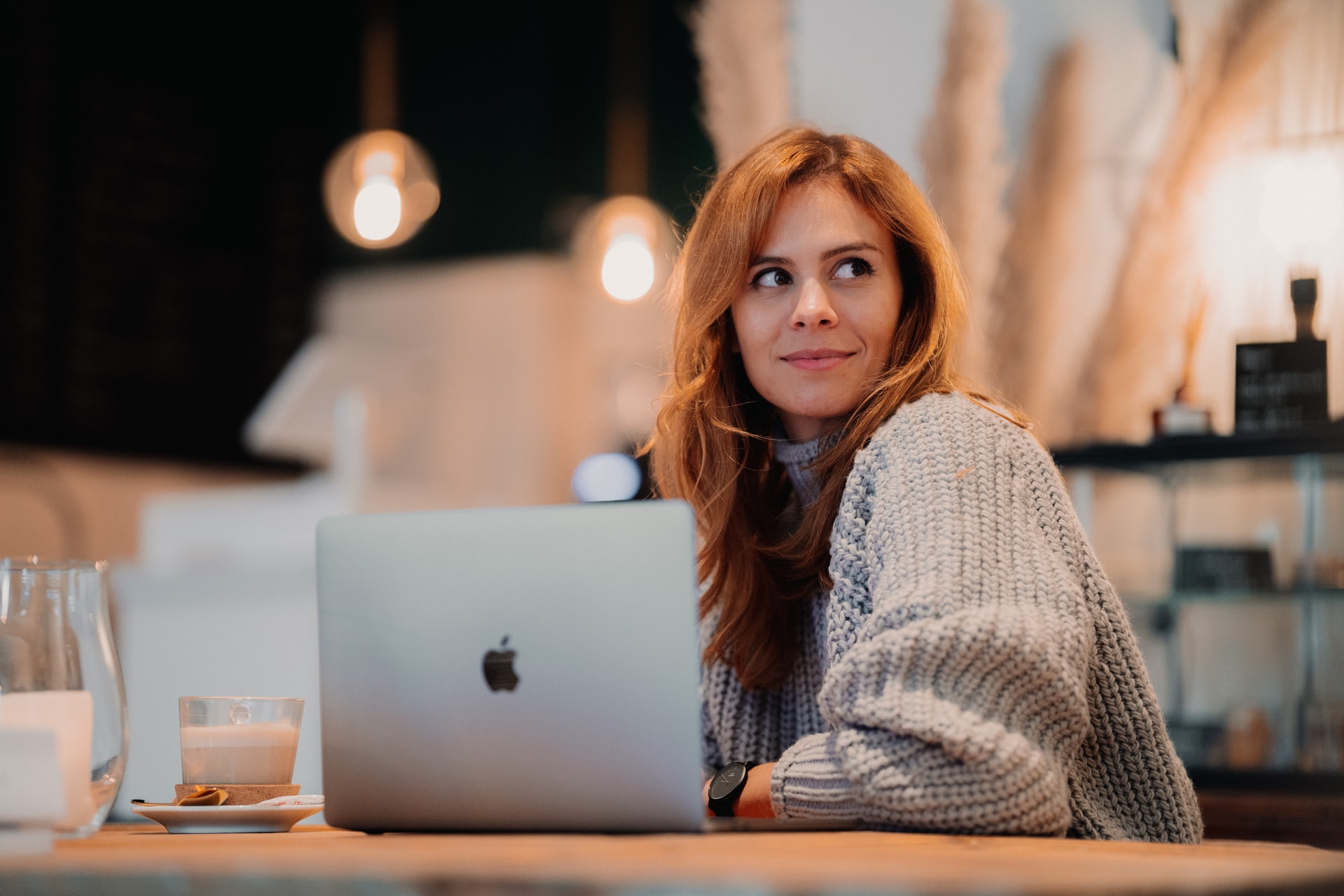 Person with red hair and grey sweater sits in a cafe, with their laptop.