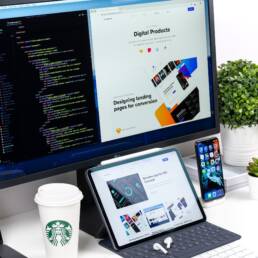 Three screens on a desk—a monitor with code, a laptop displaying a website, and an iPhone on the Home Screen.