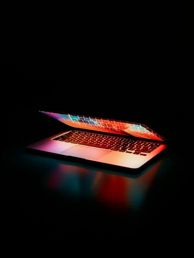 A semi-shut laptop reflecting colors on a black backdrop.