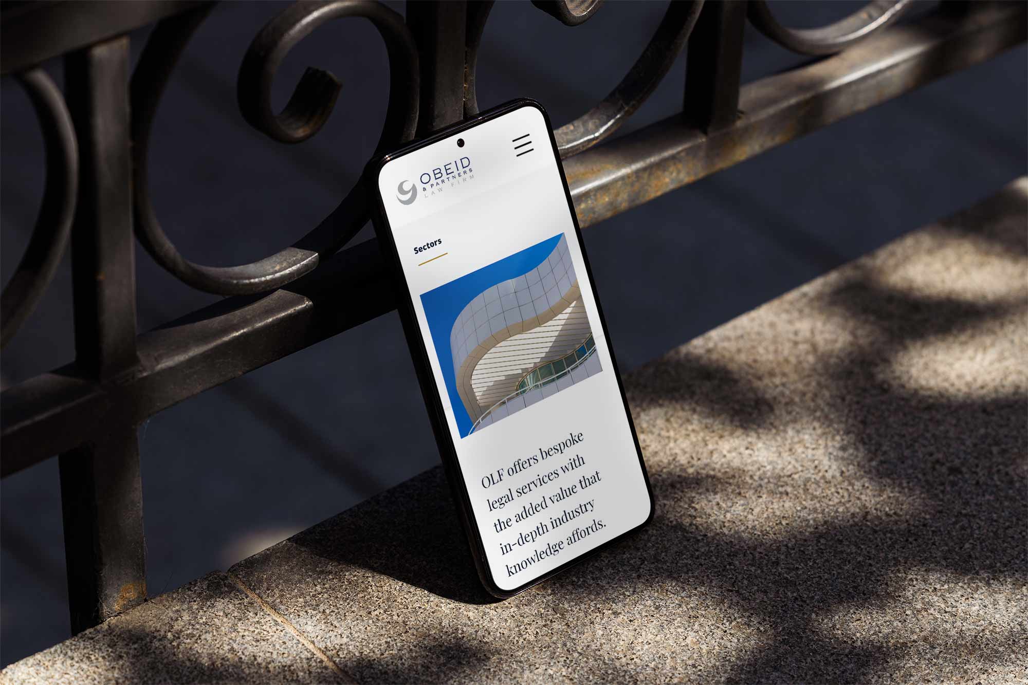 A smartphone on the ground resting against a railing displaying a law firm website with an image of a building.