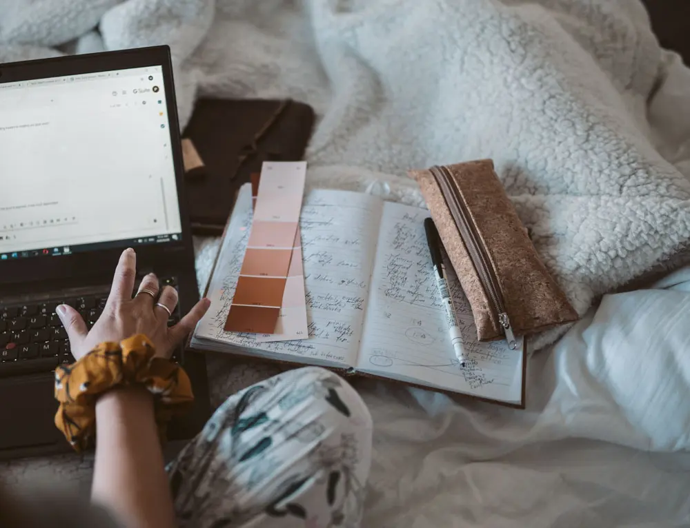 A person uses a laptop on a bed with swatches on its side.