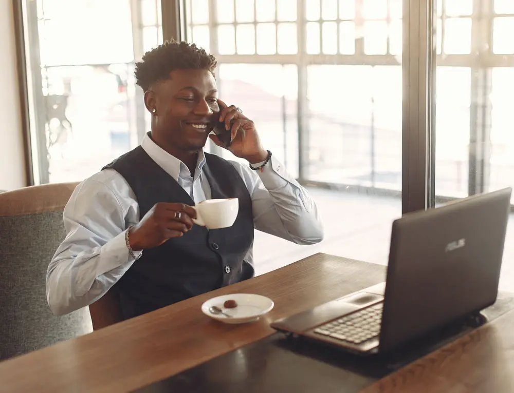 A person enjoys a coffee while talking on a phone in front of a laptop.