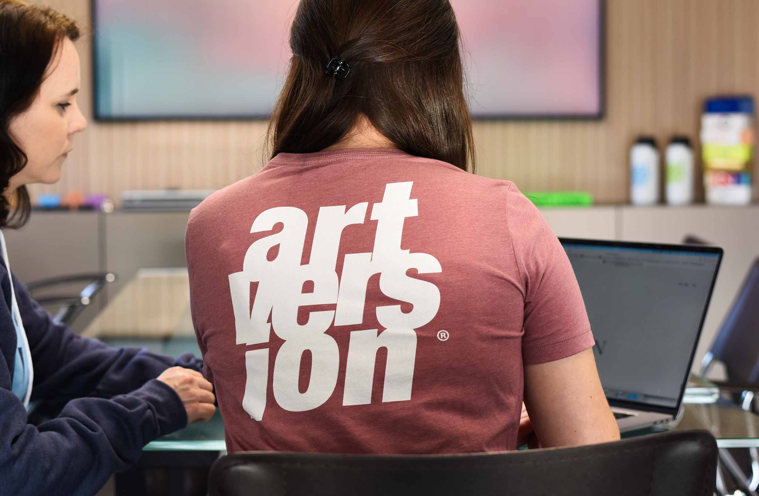 ArtVersion team member in rose t-shirt working on laptop while being observed by a colleague